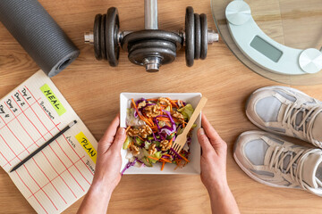 Woman holding a vibrant, healthy salad while surrounded by dumbbells, a fitness mat, a scale, and a...