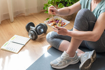 Sitting on a yoga mat, a mature woman enjoys a healthy salad after her home workout, demonstrating...