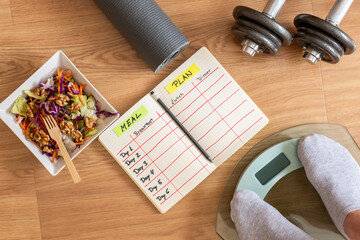 Healthy lifestyle concept with a woman weighing herself on a scale, a meal plan notebook, a bowl of salad, dumbbells, and a yoga mat, promoting balanced nutrition and exercise