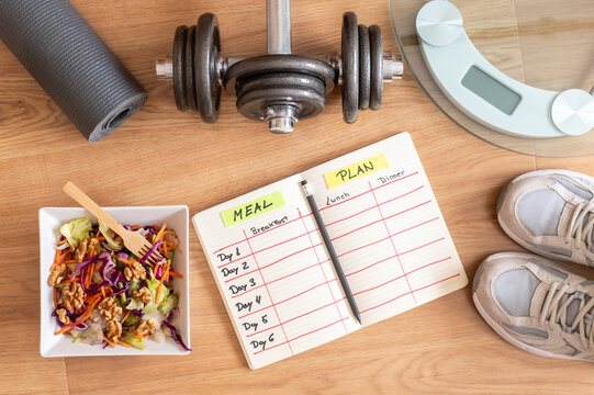 Bird's-eye view of a wooden floor featuring a healthy salad, meal planning notebook, dumbbell, weight scale, yoga mat, and sneakers, embodying fitness and balanced nutrition