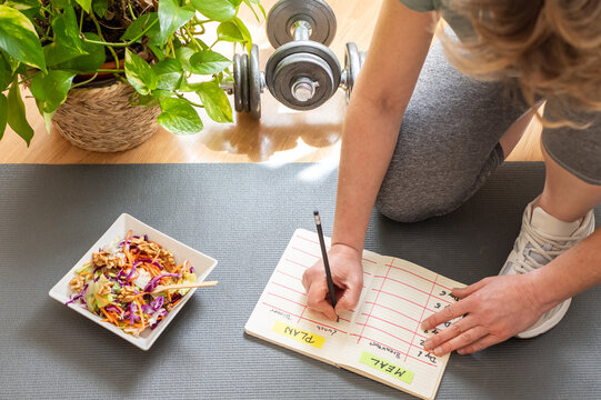 Kneeling on a fitness mat, a woman writes down her dietary plan in a notebook, next to a healthy mixed salad and a dumbbell, suggesting a focus on post-workout nutrition