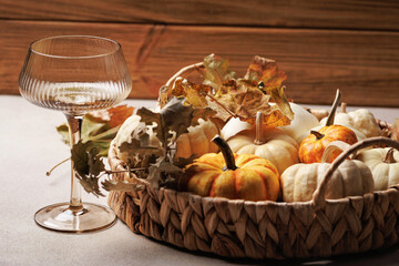 Wicker basket filled with small pumpkins and dry leaves next to an elegant wine glass on a neutral table