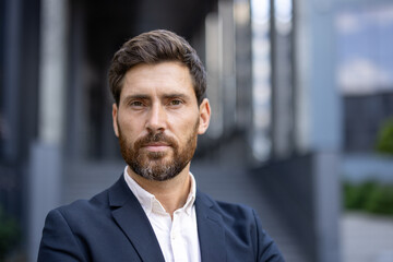 Fototapeta premium A handsome, bearded man in a suit poses confidently, looking directly at the camera with a composed expression. The urban backdrop hints at a professional setting.