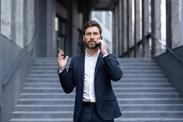 A businessman in a suit gestures while speaking on his phone, standing near stairs outdoors. The...