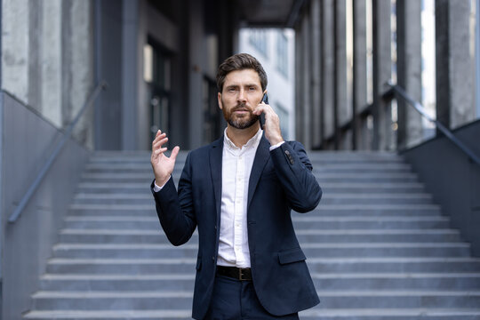 A businessman in a suit gestures while speaking on his phone, standing near stairs outdoors. The urban setting provides a modern, professional backdrop for the communication.