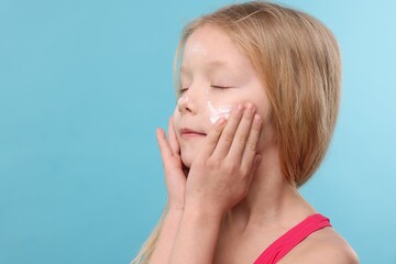 Cute little girl applying sun protection cream onto her face against light blue background, closeup. Space for text