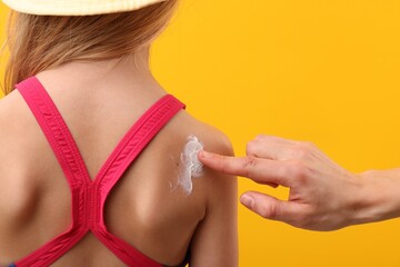 Mother applying sun protection cream onto her daughter's skin on yellow background, closeup