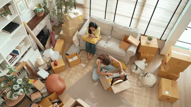 Top down view of friend unpacking box while roommate playing smart phone on sofa. Attractive woman checking furniture at package and preparing for moving while the other using social media. Myrmidon.