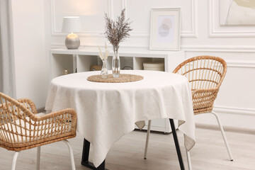 Table with white tablecloth, dried flowers in vases and chairs in stylish room. Interior design