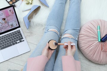 Blogger holding face powder and brush near laptop with beauty blog web site on floor indoors, above...