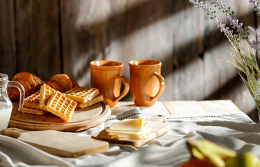 Cozy morning breakfast with waffles, croissant and milk and hot coffee on rustic wooden table. Warm sun light on wall background.