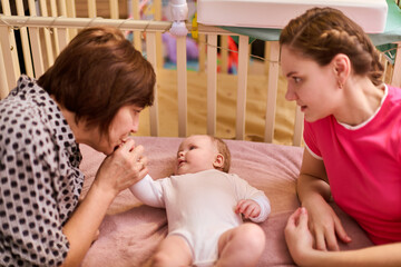 Elderly grandmother with short brown hair and young brown-haired mother watch smiling baby girl in...