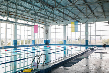 Indoor five-lane swimming pool with clear water, lane markers, and a ladder for entry. A smooth surface of water in which the interior is reflected. Large windows provide natural light.
