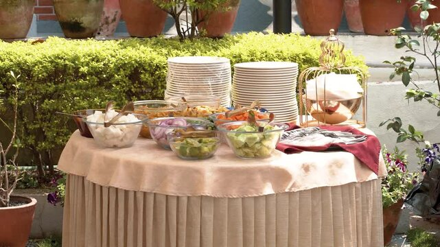 Buffet table with salads, appetizers, plates, and a covered dish, set against a green hedge for an outdoor event.