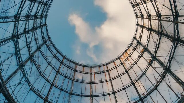 Looking up through an abandoned gasometer framework towards a cloudy sky, intricate rusted metal grid with clear blue sky visible above.