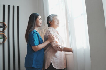 An elderly Asian woman stands at her bedroom window looking and thinking about things and an Asian nurse is taking care of her in a nursing home in the morning.