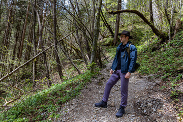 Naklejka premium A young man of about thirty in a wide-brimmed hiking hat and clothes, with a backpack, stands on a picturesque Alpine path, in the spring in Austria