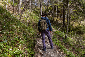 Obraz premium A young man in his thirties wearing a wide-brimmed hiking hat and clothes, with a backpack, walks along a picturesque alpine path, in spring in Austria