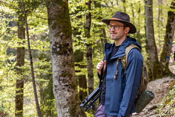 A young man in his thirties wearing a wide-brimmed hiking hat and clothes, with a backpack and a photo tripod, stands on a picturesque alpine path in spring in Austria