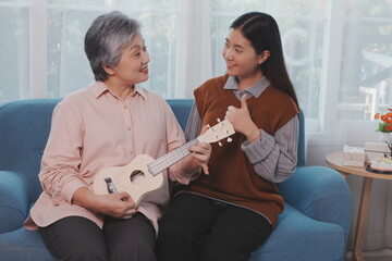 Asian elderly mother and her beautiful 29-year-old daughter relax on the sofa playing ukulele happily together at home.