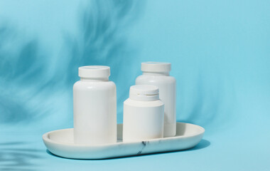 Three white plastic pill bottles of varying sizes stand on a white marble tray against a light blue background with subtle leaf shadows