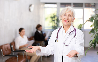 Smiling friendly elderly female doctor in white coat making welcome gesture, politely inviting...