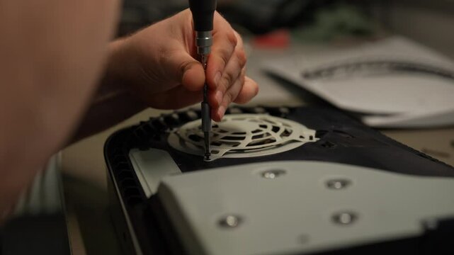 Close-up of technician using screwdriver to fix internal cooling fan of video game console, showcasing intricate process of electronic repair in workshop environment. Shooting in slow motion.