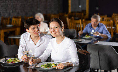 Smiling young guy spending time with adult woman in restaurant. Carefree couple laughing merrily, enjoying delicious food and drinking wine at dinner. Age-gap relationships concept