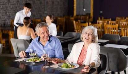 Elderly couple of spouses, husband and wife, communicate in restaurant during dinner. Mature man and woman have date in cafe, eat Oriental cuisine, drink wine tasting