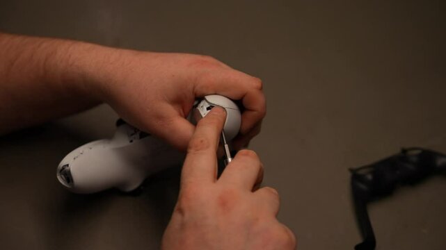 Male hands dismantling white gaming controller at table during repair process in workshop. Technician repairing broken joystick on workbench, using screwdriver to disassemble device, slow motion.