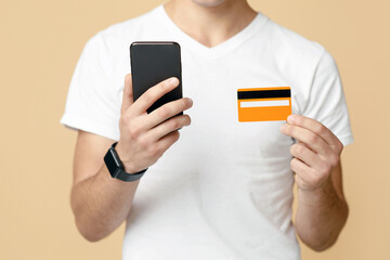 Cropped of man in white t-shirt holding smartphone and credit card in his hands, isolated on studio background. Contactless shopping, ecommerce