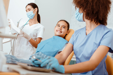 Fototapeta premium Work of modern dentist orthodontist with children. Millennial friendly african american doctor and european nurse in uniforms, protective masks and rubber gloves start examining little girl, cropped