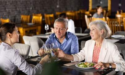 Senior parents enjoying delicious food and pleasant conversation with adult children over dinner in restaurant. Happy carefree people gathered around table for family celebration..