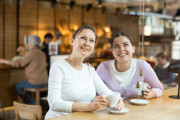 Two happy smiling young female friends having good time with cup of coffee and cheerful conversation at table at cafe