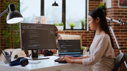 IT specialist types code on dual monitors in a cozy home office while working remotely. Asian woman uses programming language for productivity in coding and internet security. Camera B.