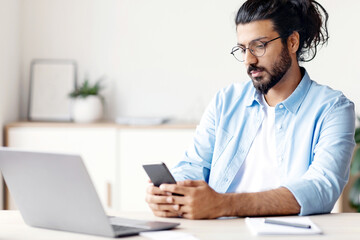 Pensive Arab Freelancer Man Messaging On Smartphone While Sitting At Desk In Home Office, Having Break In Work, Young Eastern Guy Using Cellphone, Browsing Internet Or Checking Social Networks