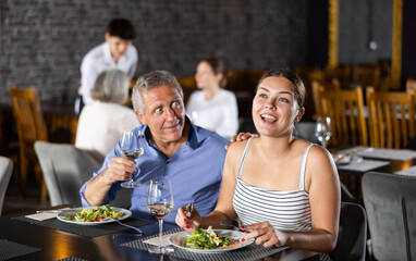 Elderly man resting in restaurant with young female companion. Couple eats dishes of national Mediterranean cuisine, drink wine, talk, communicate, laugh, joke