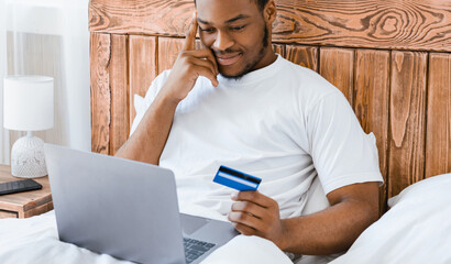 Online Shopping. Black Man With Laptop And Credit Card Making Order Sitting In Bed In Bedroom At Home. E-Commerce, Happy Customer Buying Things In Internet Shop Concept. Selective Focus