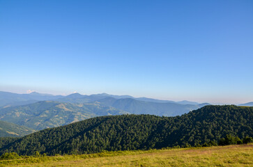 Beautiful view of a lush green mountain range under a clear blue sky with sunlight. Tranquility of nature with rolling hills, forested areas, and distant horizons. Carpathian Mountains, Ukraine
