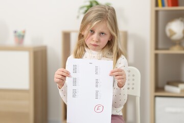 School grade. Sad girl holding answer sheet with letter F indoors