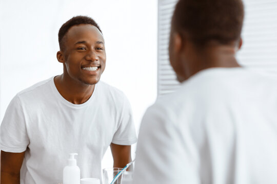 Toothcare. Happy Black Man Smiling Looking At His Healthy White Teeth In Mirror Standing In Modern Bathroom At Home. Oral Hygiene Routine And Teeth Health Concept. Selective Focus