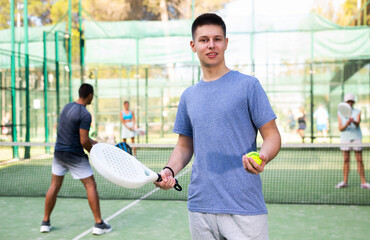 Portrait of positive caucasian boy standing on padel tennis court, holding racket and ball.