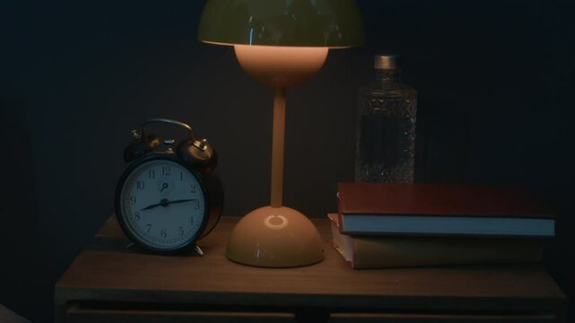 Wooden bedside table with classic vintage alarm clock, stacked books, glass water bottle and glowing lamp in dark bedroom during nighttime