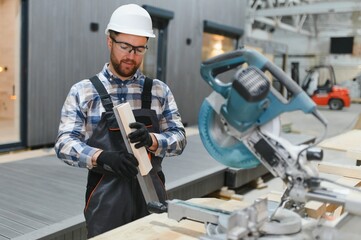A carpenter cuts a wooden plank on a sawmill in a modular building factory, providing precise cuts...