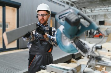 A carpenter cuts a wooden plank on a sawmill in a modular building factory, providing precise cuts...