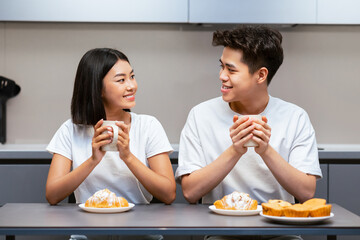 Lovely Asian Couple Having Breakfast Sitting In Kitchen, Enjoying Coffee And Croissants On Weekend...