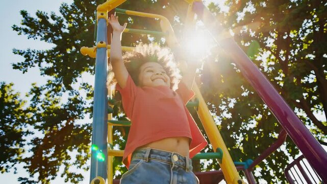 A child with curly hair in a red shirt and jeans hangs from colorful monkey bars at a playground. Trees and sunlight in the background create a lively outdoor setting.