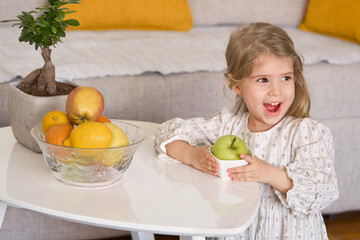 Obraz premium Beautiful little smiling girl holding fresh organic apple indoors. Healthy eating concept