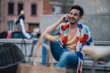 Young man talking on phone while sitting on stairs with bicycle and paper bag