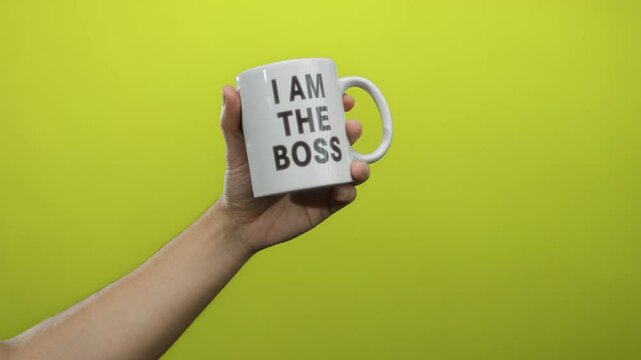 Male hand holding white mug with 'i am the boss' text against a bright yellow background isolated from other elements.